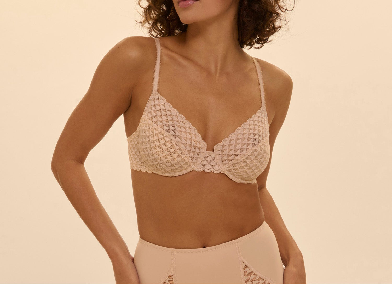 Woman wearing a beige lace bralette against a beige background