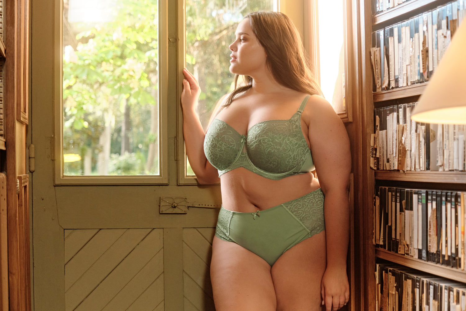 A full figured, plus sized woman stands in a library looking out the window. She is wearing a sage green bra and panty set.