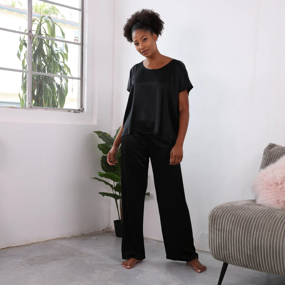 A black woman stands in a living room. She is wearing a minimalistic pajama set made of black satin. The top has a scoop neck and short sleeves. The bottoms are full length pants.