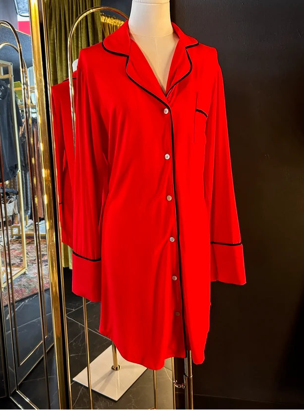 A bright red long-sleeve pajama shirt with black piping trim, displayed on a white mannequin torso in front of a gold-framed mirror against a dark wall.