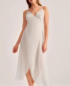 A woman modeling a white slip dress with a lace V-neck bodice and flowing chiffon skirt against a plain light background.
