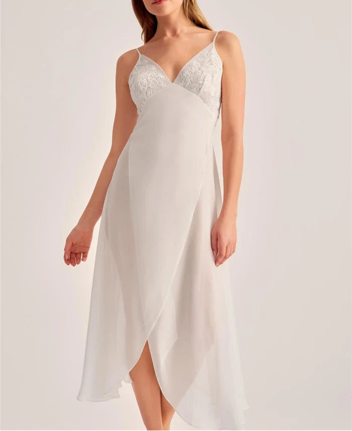 A woman modeling a white slip dress with a lace V-neck bodice and flowing chiffon skirt against a plain light background.