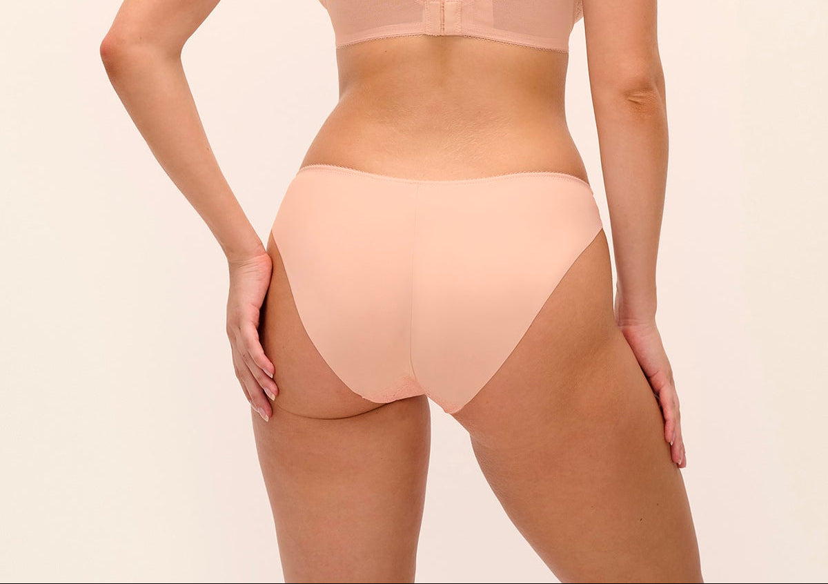 A woman shown from the back wearing a light peach-beige bra and matching underwear set against a plain light beige studio background.