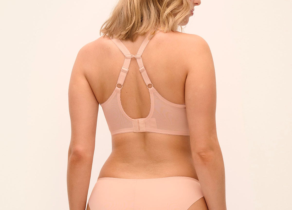 A woman shown from the back wearing a light peach-beige bralette with crossover straps and matching high-cut bikini-style bottoms, against a plain light beige studio background.