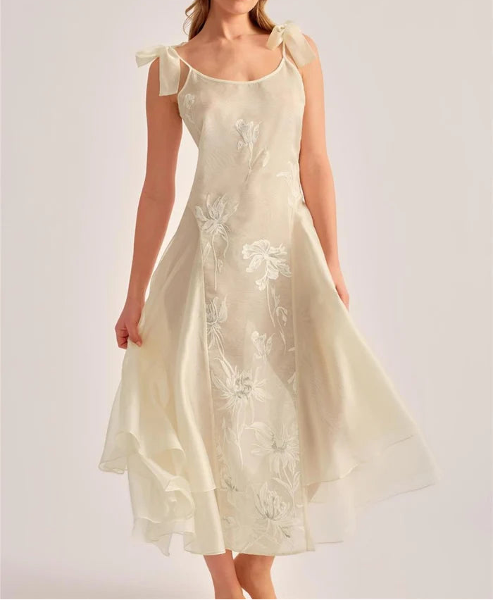 A woman modeling a cream-colored midi dress with delicate floral embroidery and sheer ruffled shoulder straps, set against a solid light gray studio background.