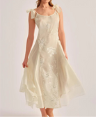 A woman modeling a cream-colored midi dress with delicate floral embroidery and sheer ruffled shoulder straps, set against a solid light gray studio background.