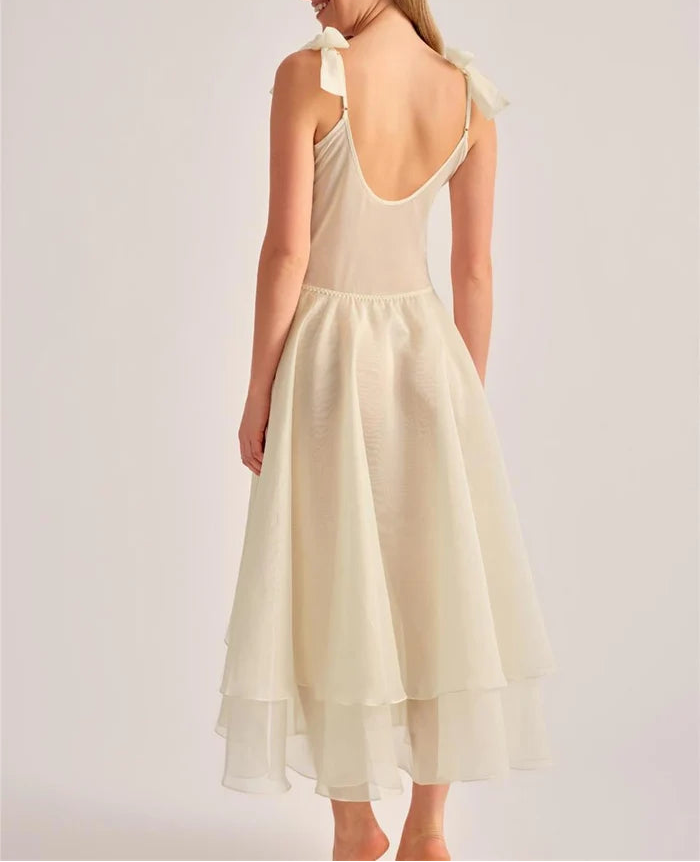 A woman shown from the back wearing a sleeveless cream dress with a deep open back, shoulder tie details, and a layered midi skirt, photographed against a plain light gray studio background.