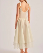 A woman shown from the back wearing a sleeveless cream dress with a deep open back, shoulder tie details, and a layered midi skirt, photographed against a plain light gray studio background.