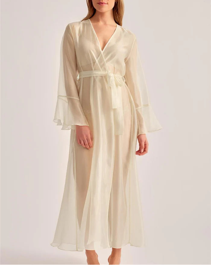 A woman modeling a long cream satin robe with a V-neck, long flared sleeves, and a matching waist tie, set against a plain light gray studio background.