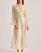 A woman modeling a long cream satin robe with a V-neck, long flared sleeves, and a matching waist tie, set against a plain light gray studio background.