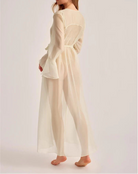 A woman modeling a long-sleeved, belted sheer maxi dress in a warm cream or off-white color, shown from a side-back angle against a plain light gray studio background.