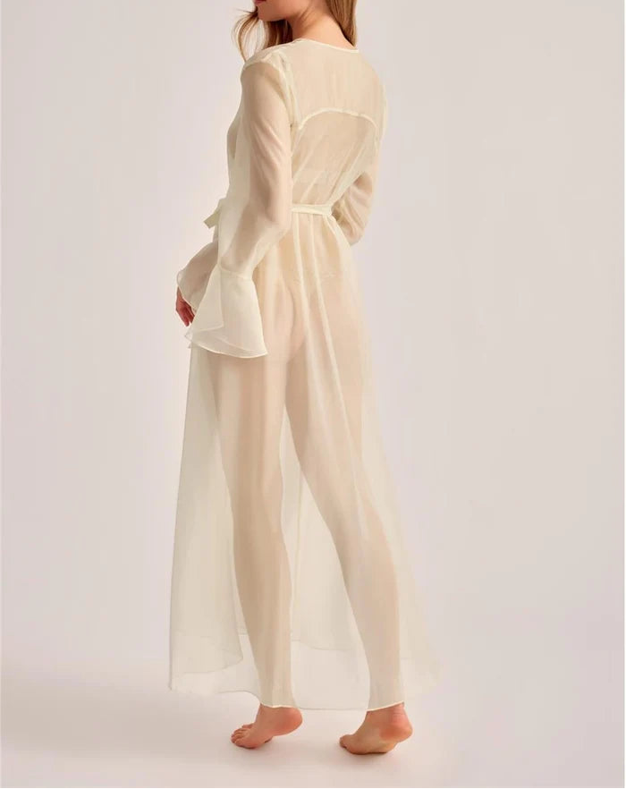 A woman modeling a long-sleeved, belted sheer maxi dress in a warm cream or off-white color, shown from a side-back angle against a plain light gray studio background.